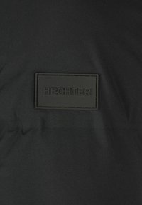 Schwarzer Stoff mit glatter Textur und einem rechteckigen schwarzen Lederetikett, das mit "HECHTER" geprägt ist und an der Vorderseite angebracht ist.