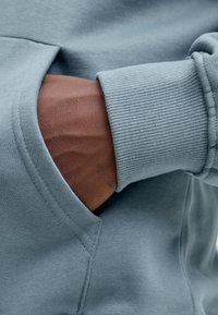 Világoskék kapucnis pulóver anyag bordázott mandzsettákkal, oldalzsebbe helyezett kézzel, sima, puha textúrát bemutatva.