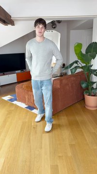 Siva majica, svetlo modre jeans hlače in beli supergi, ki jih nosi oseba, ki stoji na lesenem tleh. V ozadju sta rjav kordurov kavč in velika zelena rastlina.