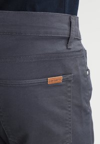 Grå bukser med en glat tekstur, der har et læder Carhartt-label på baglommen, samt subtile stikningsdetaljer.