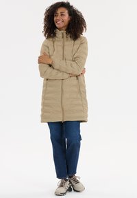 Smilende kvinde med krøllet hår iført en beige lang pufferjakke, blå jeans og beige sneakers, stående med armene korslagt.