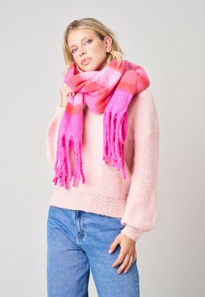 Femme aux cheveux blonds porte une épaisse écharpe rose et rouge par-dessus un pull rose clair et un jean bleu, debout devant un fond uni.