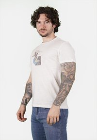 T-shirt in cotone beige chiaro con un design grafico caratterizzato da forme e texture astratte. Maniche corte e collo rotondo.