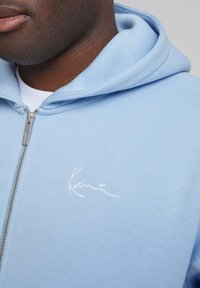 Lyseblå hettegenser med glidelås laget av myk stoff, med en hvit brodert logo på venstre bryst og snor i hetten.