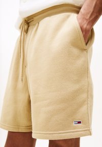 Beżowe spodenki związywane sznurkiem, z bocznymi kieszeniami, noszone przez osobę stojącą z jedną ręką w kieszeni, z małym czerwono-biało-niebieskim logo blisko dołu.