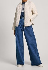 Prošívaná krémová bunda s kulatým límcem, kombinovaná s tmavě modrými džínami s širokými nohavicemi a bílými teniskami; pod ní je grafické tričko.