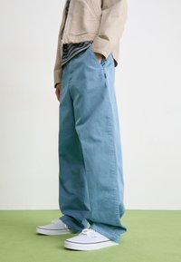 Light blue wide-leg pants made from cotton, featuring side pockets, paired with white sneakers. The background is neutral with a green floor.