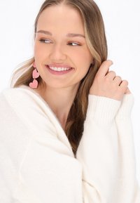 Ružové náušnice v tvare srdca visia na háčikoch a majú viacvrstvový dizajn. Modelka má na sebe jemný sveter v krémovej farbe.
