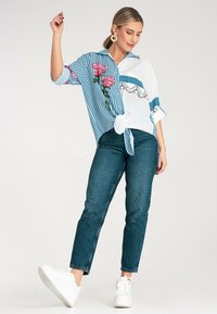 Niebieska i biała koszula w paski z kwiatowym wzorem, związana w talii, w połączeniu z ciemnoniebieskimi dżinsami i białymi trampkami. Gładkie tło.