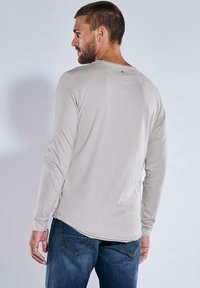 Helles graues Langarmshirt mit Rundhalsausschnitt, das mit genähten Nähten und einer leichten Saumkurve versehen ist. Getragen mit blauen Jeans.
