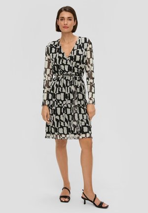 Robe noire transparente avec motif blanc abstrait, manches longues, décolleté en V profond, taille cintrée et longueur au genou. Associée à des sandales noires à talons.
