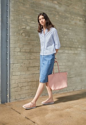 Kvinde går indendørs iført en lyseblå skjorte, denim nederdel, pink sneakers og bærer en stor pink tote taske.