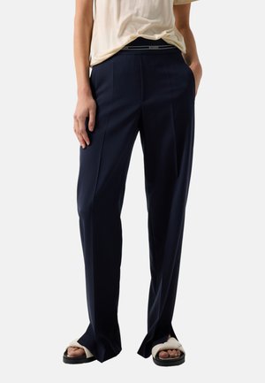 Person iført marineblå skræddersyede bukser, beige afslappede sandaler og en lysebeige skjorte stukket ned i bukserne, stående med hænderne i lommerne.