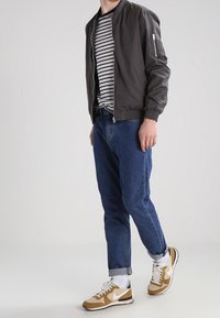 Grau Bomberjacke, gestreiftes T-Shirt, blaue Jeans und beigefarbene Nike-Sneaker mit weißen Akzenten. Die Jacke hat eine Reißverschlusstasche und gerippte Bündchen.