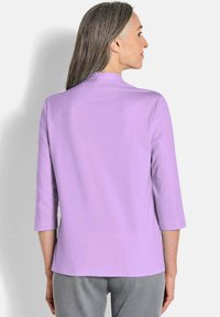 Lila Bluse mit dreiviertel Ärmeln, subtiler Struktur, rundem Ausschnitt und geradem Schnitt, kombiniert mit grauen Hosen. Keine sichtbaren Muster oder Akzente.