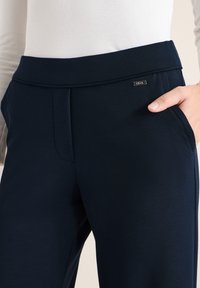 Femme portant un pantalon ajusté bleu marine foncé avec une braguette avant cachée et la main dans la poche latérale, associé à un haut crème à manches longues.