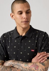 Musta polo-paita, jossa on teksturoitu kangas, esittelee geometrista kuviointia ja pieniä sinisiä ja kultaisia yksityiskohtia. Mukana on punainen Levi's-logo-tunnus.