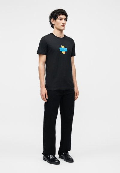 Jeune homme aux cheveux bouclés portant un t-shirt noir à motif graphique, un pantalon noir et des chaussures habillées noires, debout devant un fond clair uni.