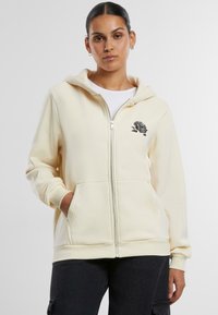 Cremefarbener Zip-Hoodie mit großen Fronttaschen und einem floral schwarzen bestickten Design auf der linken Brust. Weicher Stoff, entspannter Schnitt.