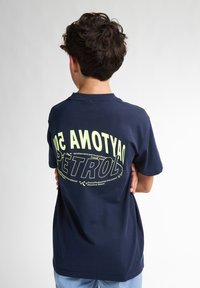Camiseta de algodón azul marino con tipografía en blanco y verde claro en la parte trasera, con un diseño circular y acentos a cuadros.