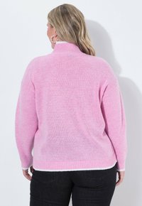 Roze gebreide trui met hoge kraag en ribbels aan de mouwen, met een zachte textuur en een licht oversized pasvorm, gedragen met donkere jeans.