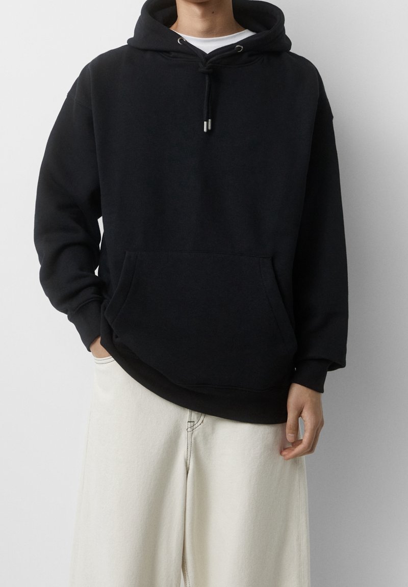 Personne portant un sweat à capuche noir avec une poche devant et des cordons blancs, associé à un pantalon blanc ample sur un fond uni.