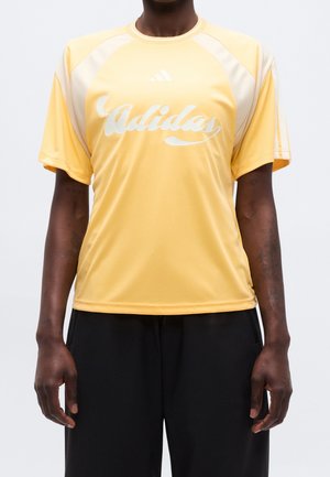 Person iført en gul Adidas t-shirt med hvidt logo og detaljer, kombineret med sorte bukser, vist fra skuldre til hofter.