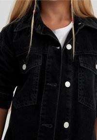 Chaqueta de mezclilla negra con cierre de botones en la parte delantera, dos bolsillos en el pecho y mangas enrolladas. Textura suave con accesorios de hardware plateado.