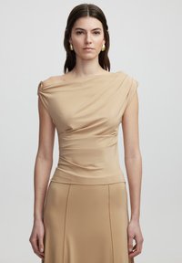 Beige off-shoulder top van soepele stof, met gedrapeerde details en een enkele decoratieve accent op de linkerschouder.