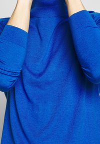 Klart blå polotröja gjord av mjukt material, med långa ärmar och ribbade manschetter samt avslappnad passform. Slät textur.