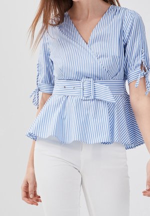 Kobieta w niebiesko-białej, pionowo paskowanej bluzce z peplum, z dopasowanym paskiem i białych spodniach, stojąca z rozluźnionymi ramionami wzdłuż ciała.