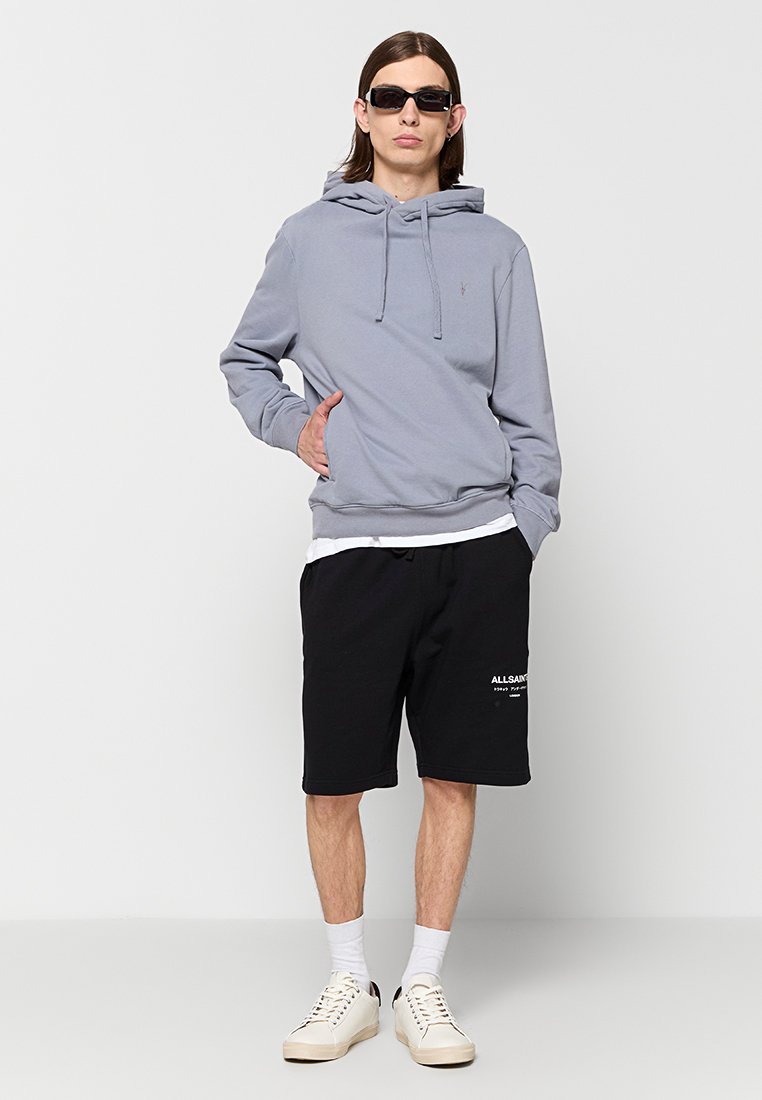 Jasnoszara bluza z kapturem i sznurkami, czarne szorty z białym logo oraz kremowe sneakersy. Model nosi okulary przeciwsłoneczne i białe skarpetki.