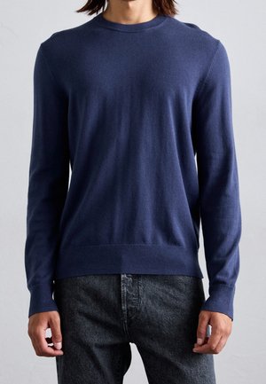 Personne portant un pull bleu marine uni à manches longues et un jean gris foncé, face à un fond uni.