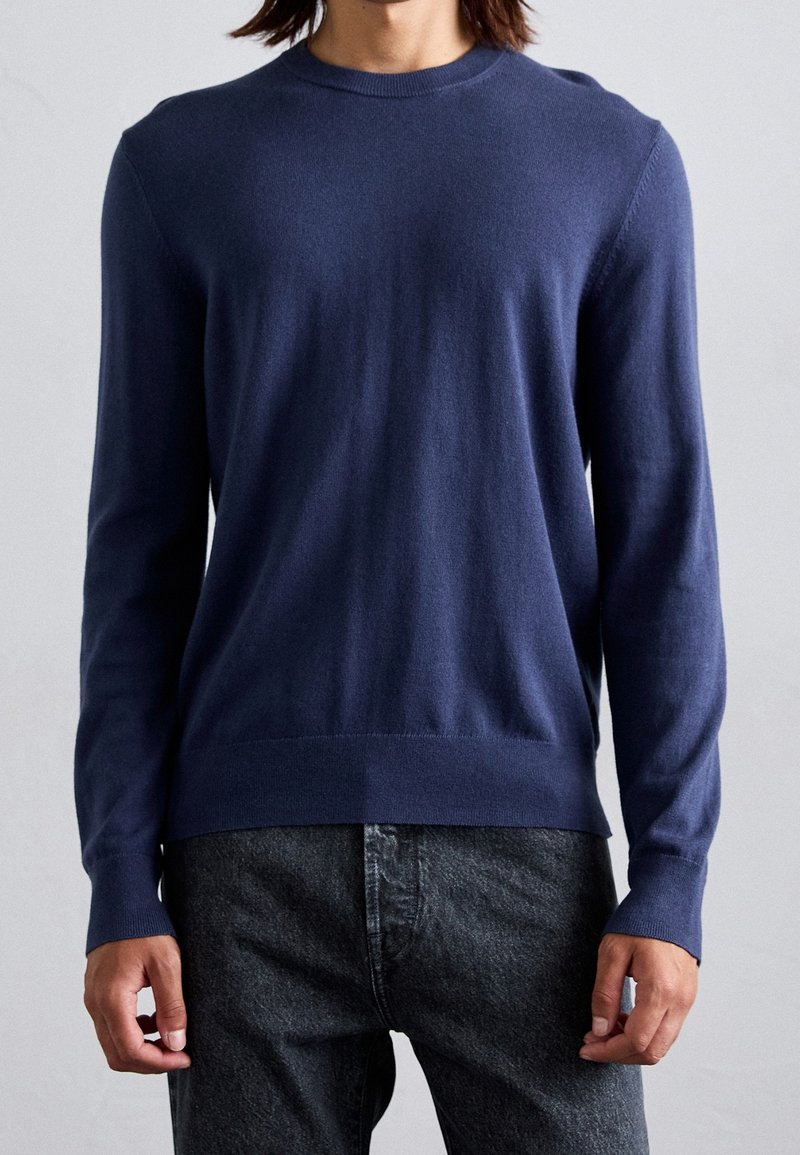 Personne portant un pull bleu marine uni à manches longues et un jean gris foncé, face à un fond uni.