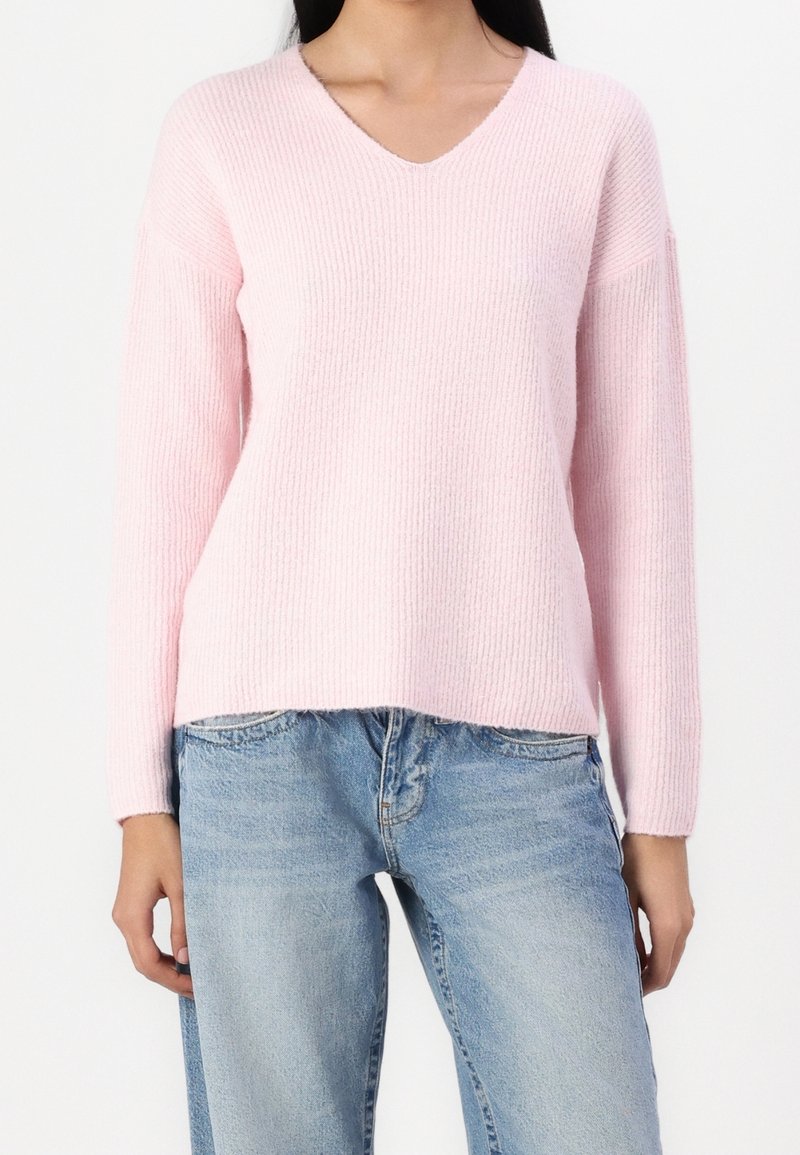 Personne portant un pull côtelé rose clair à col en V et un jean en denim bleu clair, debout sur un fond blanc uni.