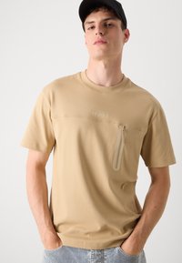 Beige színű pamut póló, elülső cipzáras zsebbel, rövid ujjakkal és logóval a mellkasán. A szabása kényelmes, kerek nyakkivágással.