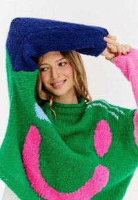 Strickpullover mit grünem Körper, rosa und blauen Akzenten und einem blauen Ärmel. Verfügt über ein Smiley-Muster und einen strukturierten Stoff.