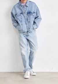 Giacca di jeans azzurro chiaro con vestibilità morbida, due tasche sul petto e chiusura con bottoni, abbinata a jeans azzurro chiaro e sneakers bianche.