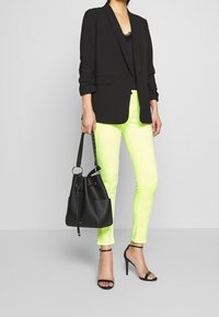 Blazer negro, jeans ajustados verde neón, bolso tipo cubo negro con acabado texturizado y sandalias de tacón alto negras con tiras.