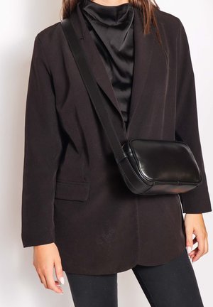 Vrouw draagt een zwarte blazer en een satijnen top met een zwarte leren crossbodytas over de schouder, staand tegen een effen achtergrond.