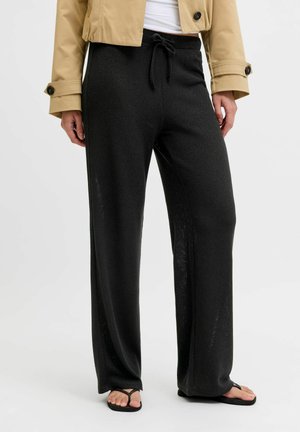 Czarne spodnie o szerokich nogawkach ze ściągaczem, noszone z beżową kurtką i czarnymi klapkami na osobie stojącej na jednolitym tle.