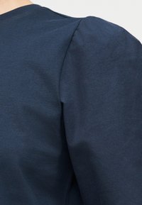 Κοντινό πλάνο ενός ατόμου που φοράει μπλε ναυτικό πουκάμισο με φουσκωτή λεπτομέρεια στους ώμους και ορατή ραφή στο πάνω μέρος του βραχίονα.