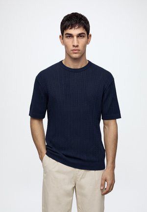 Homme portant un pull texturé bleu foncé à manches courtes et un pantalon beige clair, debout avec une main dans la poche devant un fond uni.