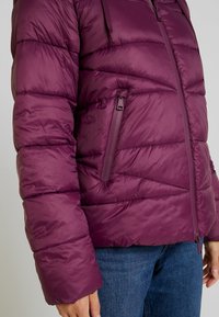 Kurtka puchowa w głębokim purpurze z poziomym pikowaniem. Posiada zamkniętą kieszeń po boku, fakturowany materiał i kaptur.