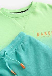 Felpa verde e pantaloni jogger blu, entrambi in morbido cotone. La felpa presenta un logo arancione; i pantaloni jogger hanno cordini azzurri chiari con punte arancioni.