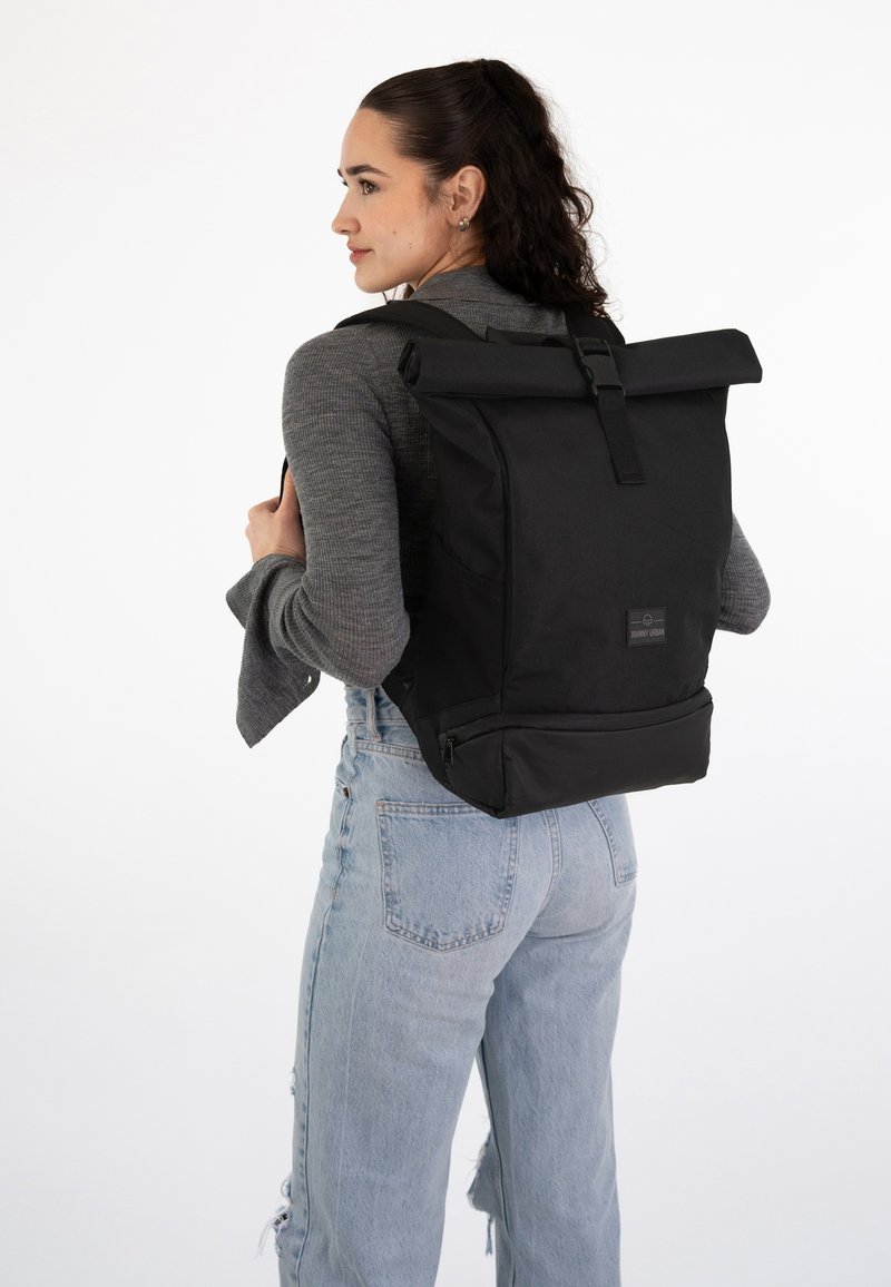 Kvinde iført en sort rulle-top rygsæk, grå langærmet top og lyseblå revnede jeans stående mod en hvid baggrund.