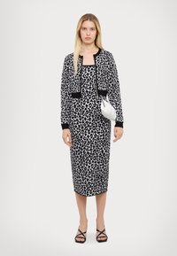 Robe à imprimé léopard avec des accents noirs, associée à un cardigan noir court. Le modèle tient un sac à main blanc brillant. Sandales à brides noires.