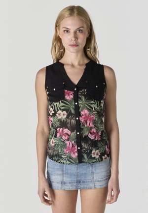 Mujer rubia con blusa negra sin mangas con estampado floral en rosa y verde y mini falda vaquera azul claro, de pie contra un fondo gris.