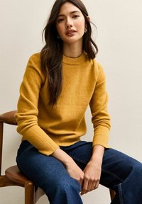 Mosterdgele gebreide trui met een geribbelde ronde hals en aansluitende manchetten, gecombineerd met donkerblauwe wijde jeans, zittend op een houten stoel.