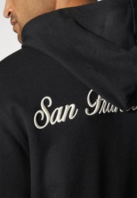 Čierna mikina s kapucňou, ktorá má jemnú textúru a na chrbte je vyšitý nápis "San Francisco" bielym písmom. Obsahuje aj šnúrku na stiahnutie kapucne.